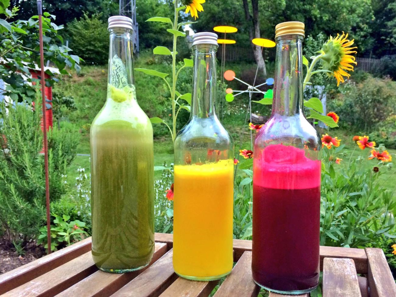 Man sieht drei Glasflaschen auf einem Tisch im Garten stehen, die mit farbigen Säften gefüllt sind